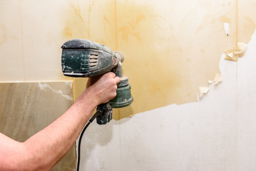 Een professionele behangverwijderaar gebruikt een plamuurmes om voorzichtig vliesbehang van de muur te verwijderen, terwijl een emmer met warm water en een spons naast hen staat.
