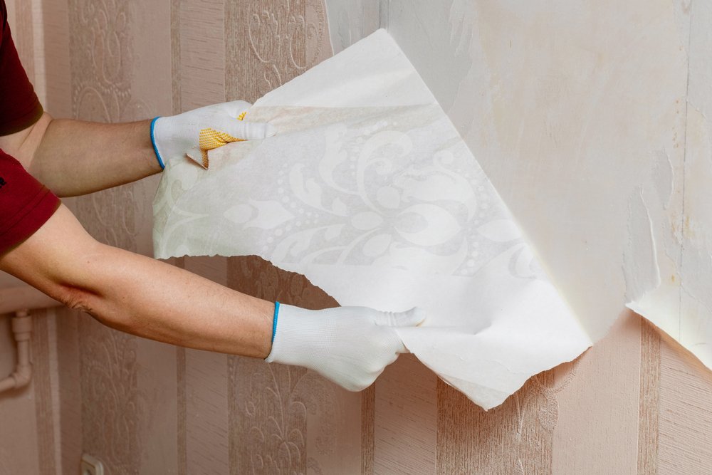 Een persoon gebruikt een plamuurmes om voorzichtig vliesbehang van de muur te verwijderen, terwijl een emmer met warm water en een spons klaarstaat voor het schoonmaken van de muur.