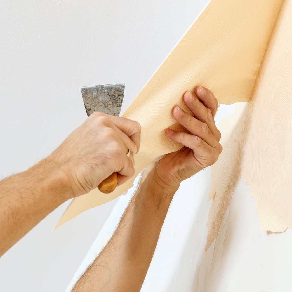 Een persoon gebruikt een plamuurmes om voorzichtig vliesbehang van de muur te verwijderen, terwijl een emmer met warm water en een spons naast hen staat.