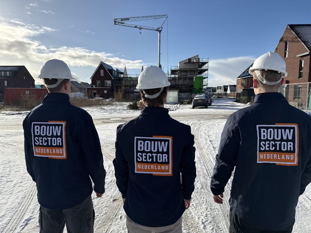 Drie vakmensen van Bouwsector Nederland met witte helm en bedrijfsjas op een besneeuwde bouwplaats met een kraan